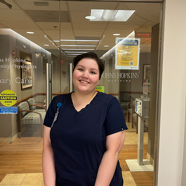 Nina Steigerwald, wearing navy blue scrubs, poses for a photo in front of doors to JHCP’s Bethesda location.