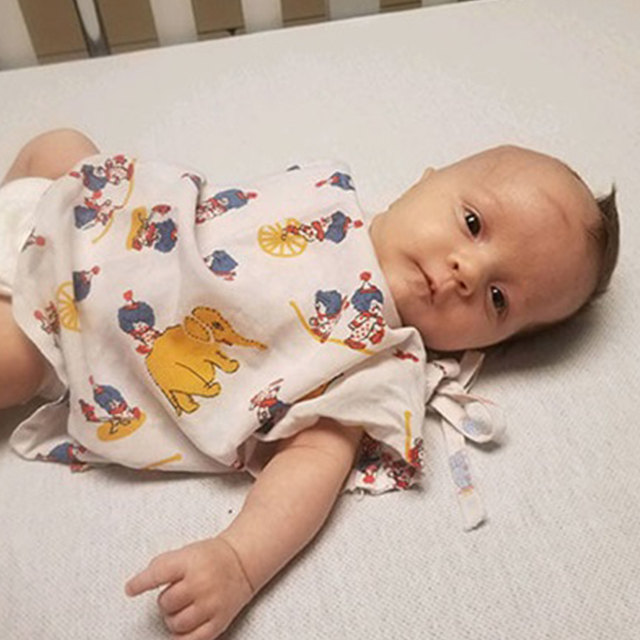 Baby Julian lays on a bed in patterned pjs. Half of his head is shaved bald.  