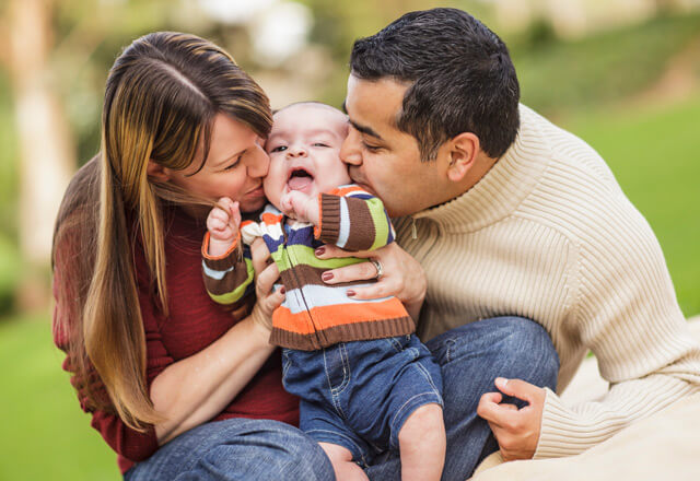 two parents kissing their infant on the cheek at the same time in a park