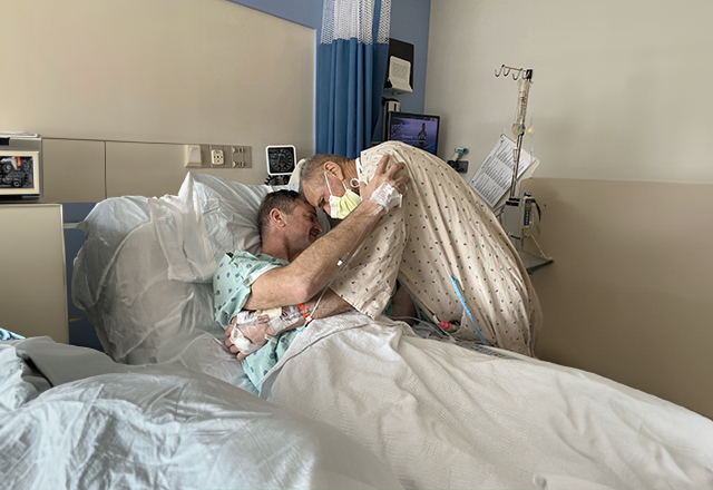 Two men in hospital gowns hugging. One in a hospital bed.