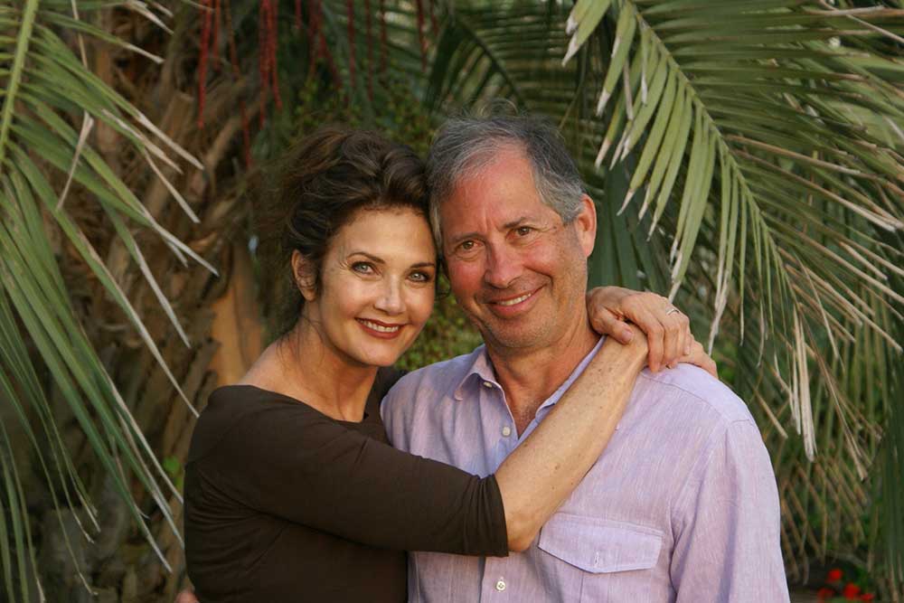 Robert and Lynda Altman smiling together in front of lush tropical foliage