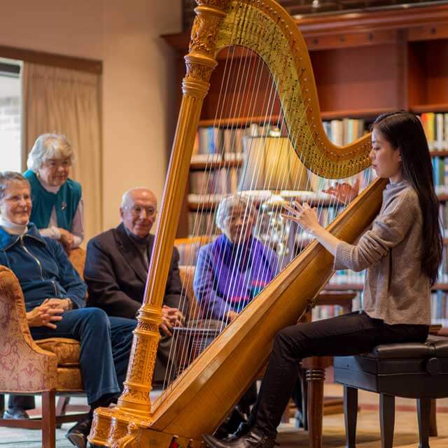 Harpist Peggy Houng playing a harp in a library in front of four older people sitting on a couch