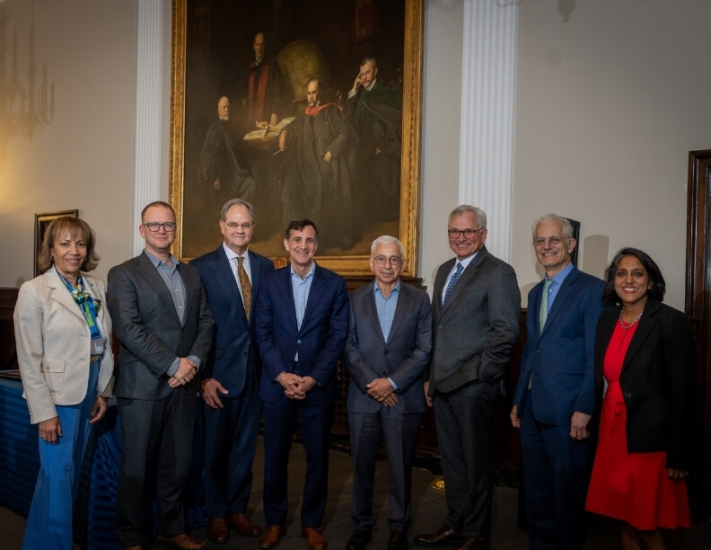 Dr. Maria Oliva-Hemker, Dr. Jeremy Greene, Dr. Landon King, JHU President Ron Daniels, Dr. Charles Homcy, Dr. Paul Auwaerter, Dr. David Thomas, and Dr. Amita Gupta pose for a photo in front of The Four Doctors painting in the Welch Library West Reading Room