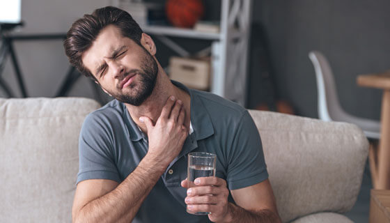 Frustrated handsome young man touching his neck and holding a glass of water while sitting on the couch at home