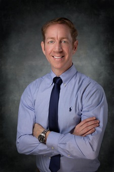 Yuval Pinto smiles for a photo with his arms crossed in front of a gray background.