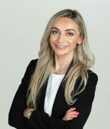 Danna Kashlan smiles for a photo in a black suit with a white top in front of an off-white background.