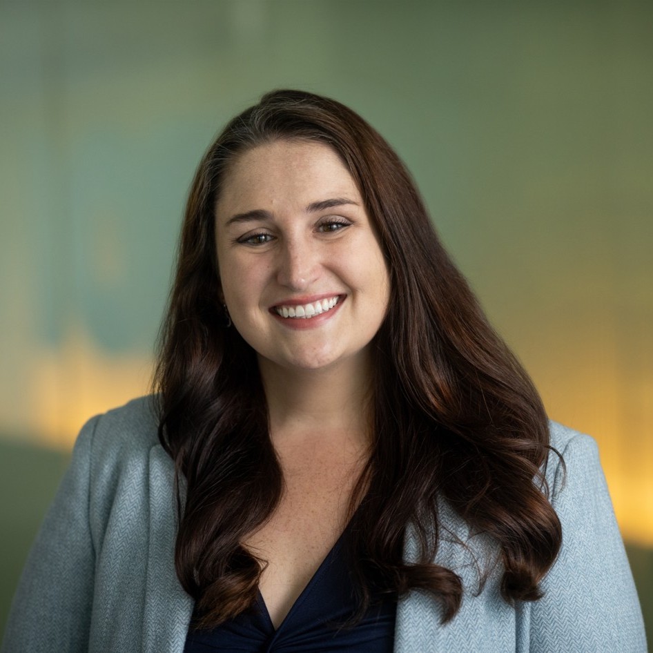Katie Caviness smiles for a photo in front of a low-lit blue/yellow background in the Johns Hopkins Bloomberg School of Public Health. Katie has long dark hair that is curled and is wearing a light blue blazer and navy top.