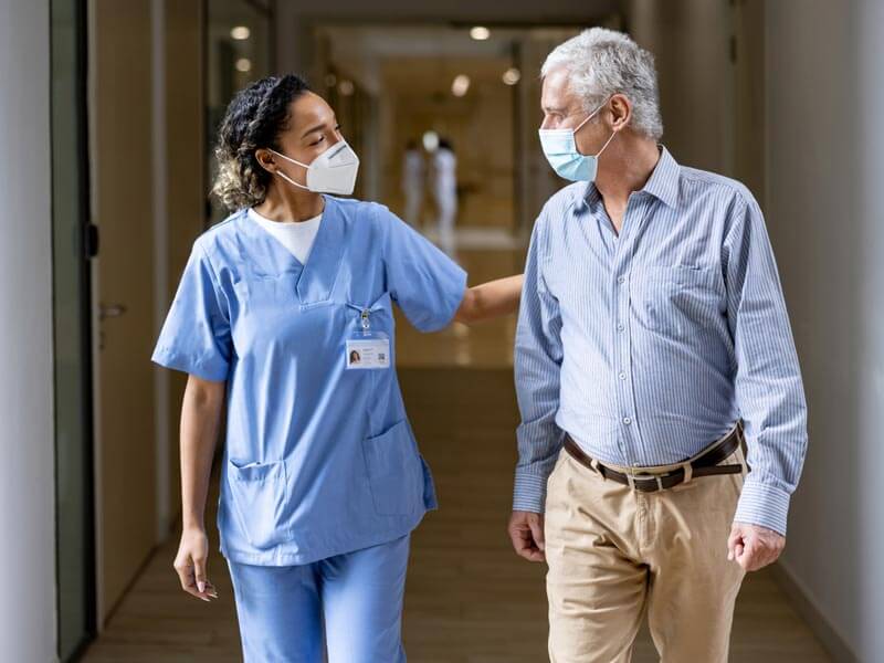 Masked doctor talking with senior patient walking down hallway