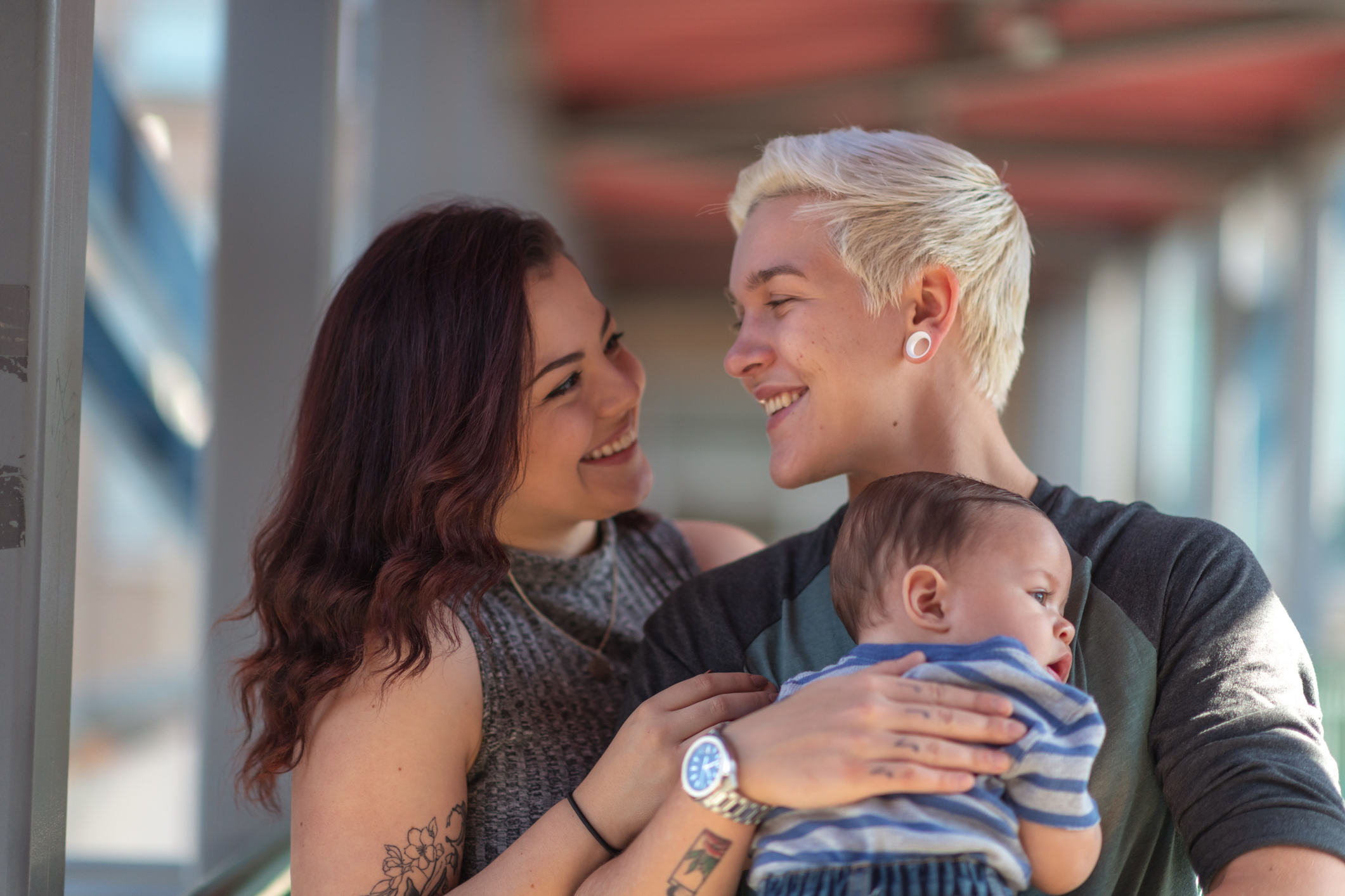 Transgender couple smiles at each other while holding baby