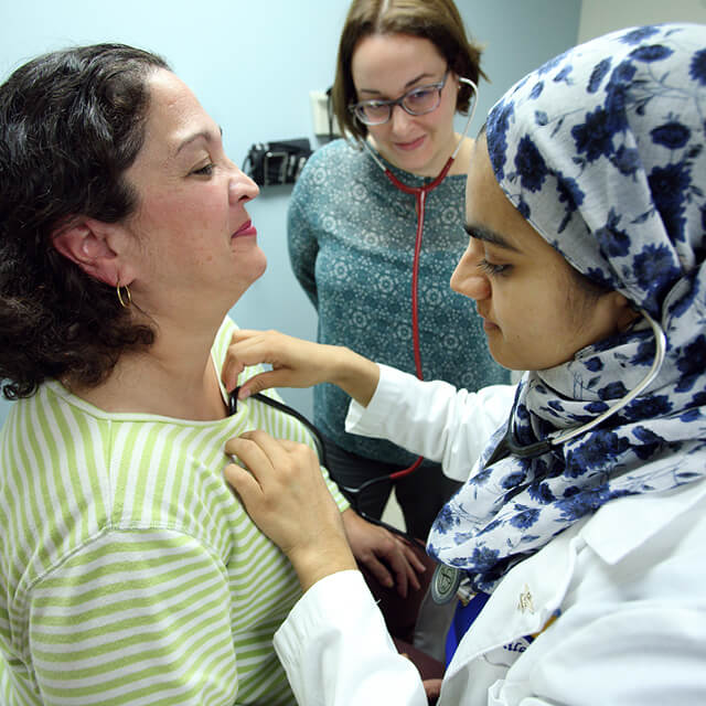 Using a shared stethoscope, med student Anila Chaudhary listens to patient Maria Aponte’s heart as internist Fernanda Porto Carreiro looks on. 