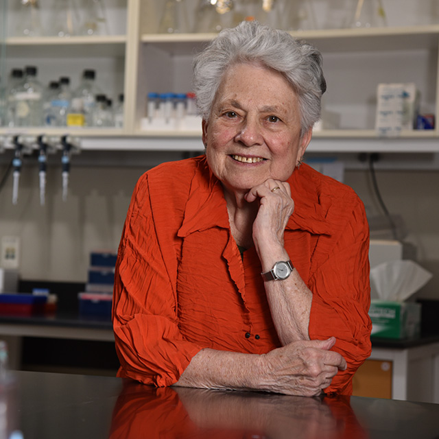 photo of Barbara Migeon, wearing a red shirt, smiling with her hand on her chin. 