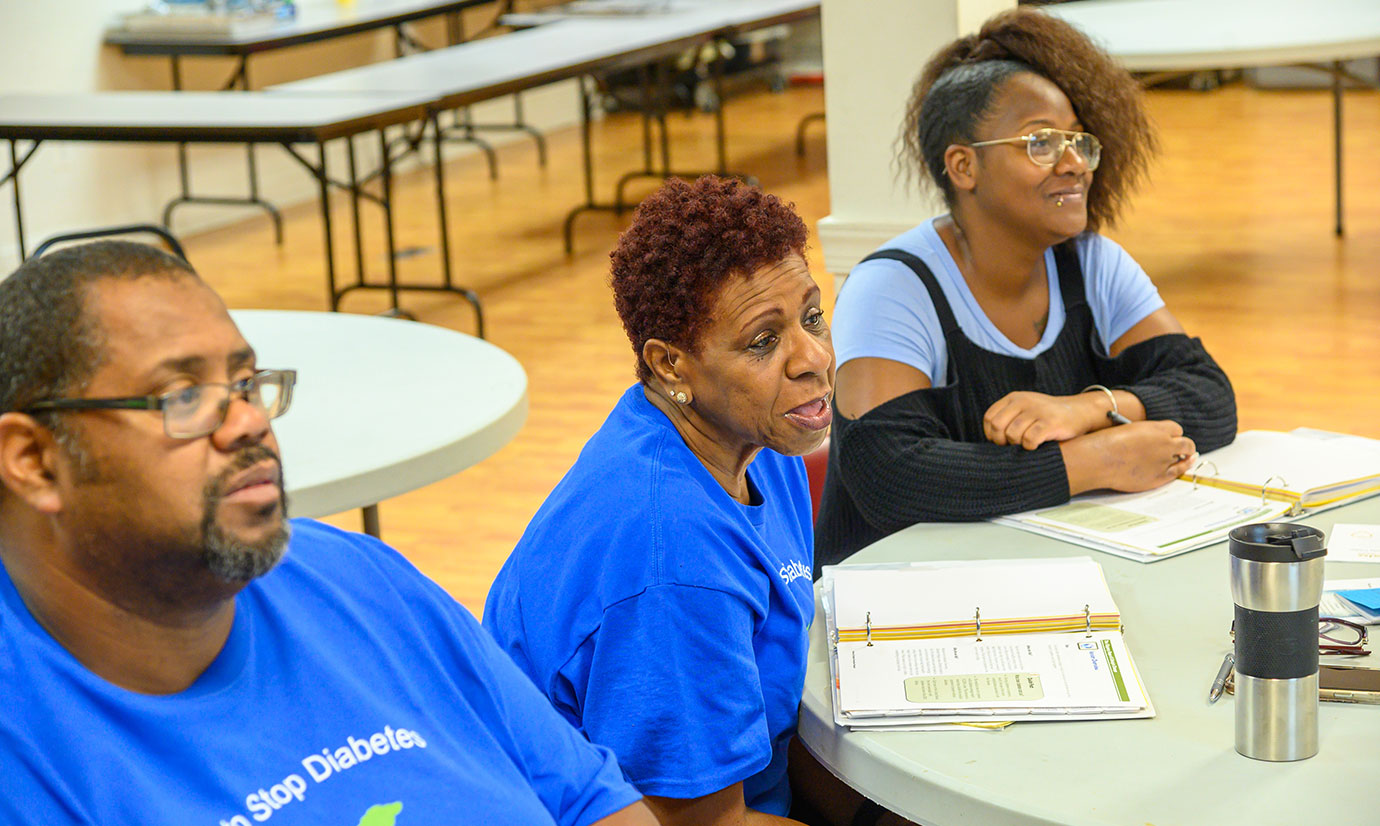 A middle-aged woman wearing a blue shirt sitting between a middle-aged man and young woman speaks at a DPP meeting.
