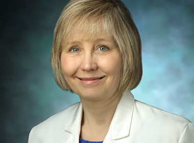 Dr. Valerie Baker wearing a traditional white lab coat with a blue blouse underneath on a blue background.