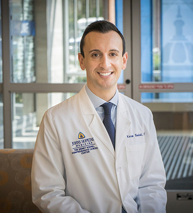 Medical oncologist Karim Boudadi wears a white lab coat, light blue button down shirt and navy blue tie in a formal portrait with a hospital entrance behind him.