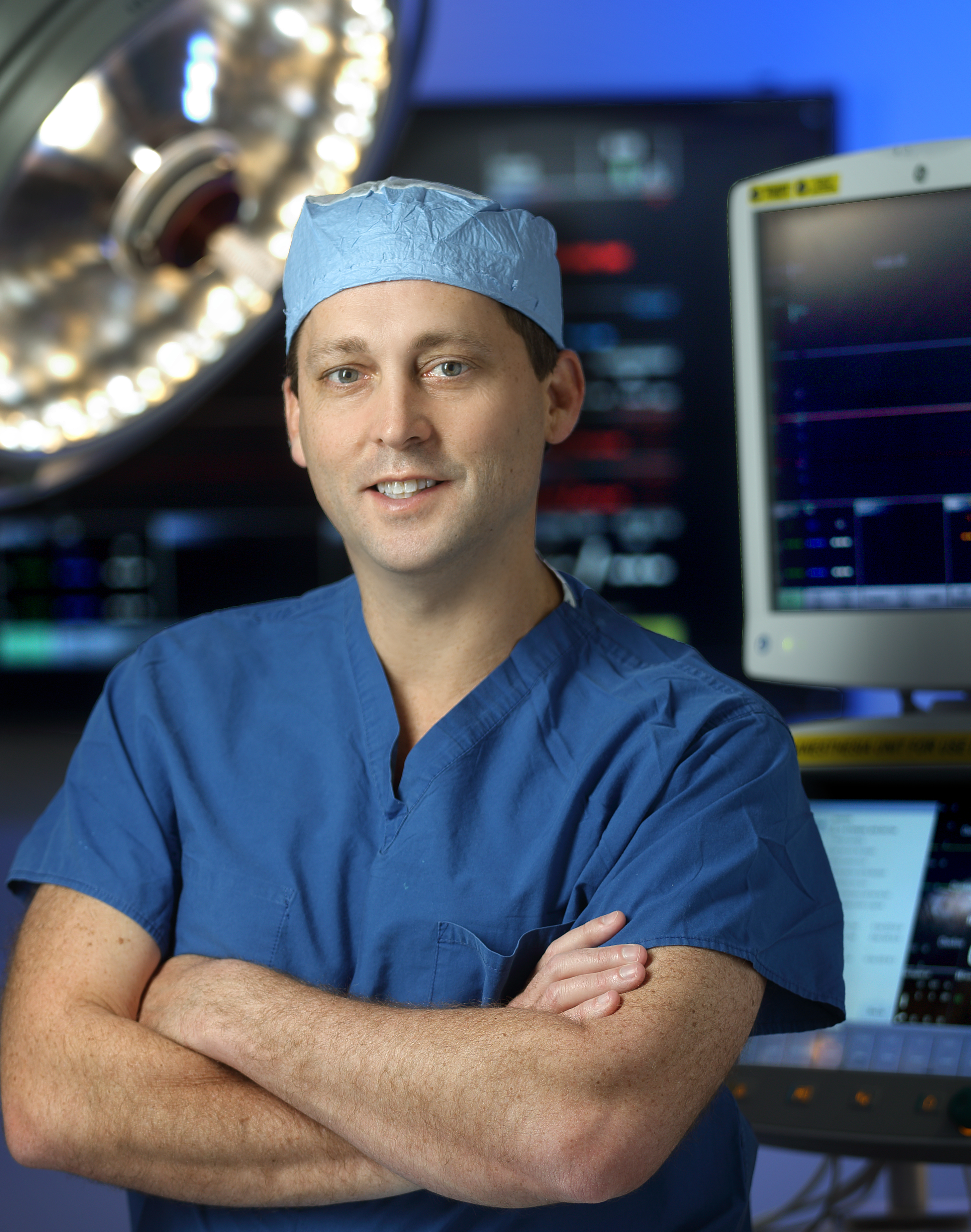 Smiling Bret Mettler stands with arms crossed in operating room while wearing blue scrubs. 