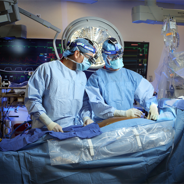 Cardiothoracic surgeon Ahmet Kilic and a surgical resident, both wearing gowns, gloves, surgical masks, surgical caps and surgical loupes, perform a surgery in an operating room.