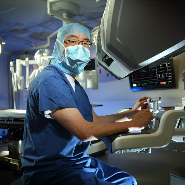 Surgical oncologist Jin He, wearing blue scrubs, a head covering and face mask, sits with his hands on the controls of a surgical robot. 