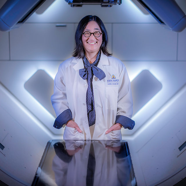 : Christina Tsien, wearing a white lab coat, inside the proton center with an apparatus above her head and rounded white walls behind her.