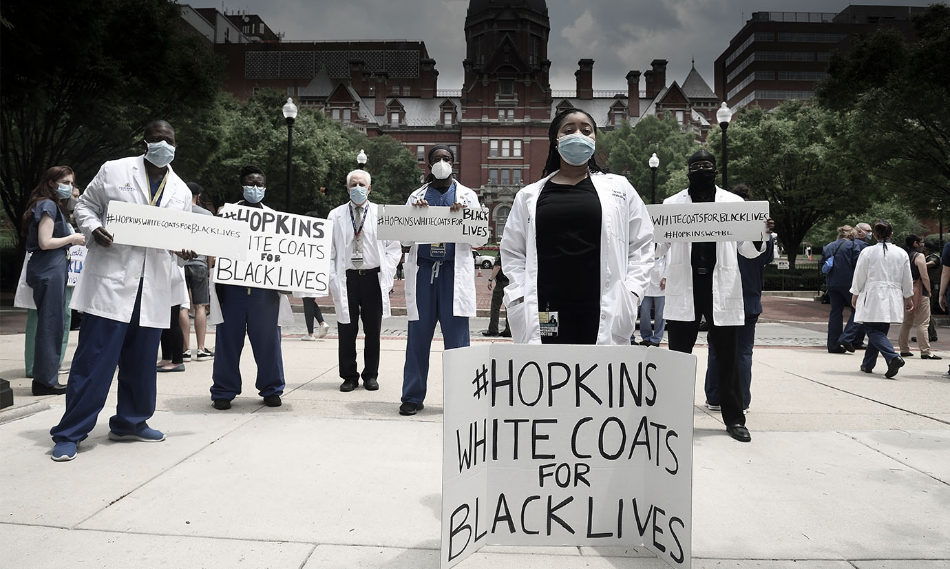 A photo of Doctors and Nurses standing in response to the Black Lives Matter movement. In front on the primary woman, there is a sign that reads, "Black Lives Matter"