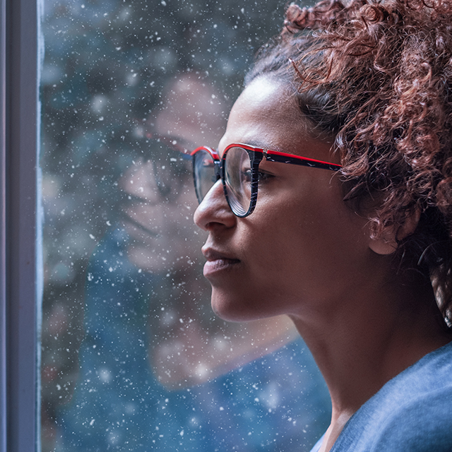 Photo of an African American woman in a blue tee shirt staring, sadly, out a window. Outside the window it's snowing. 