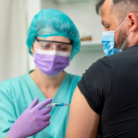 A woman wearing a light blue hairnet and scrubs, purple gloves and mask, and clear protective glasses inserts a needle into a man's upper arm. The man is wearing a black t shirt and light blue mask.