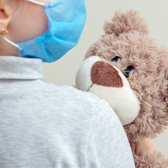 A photo shows a child wearing a mask and holding a teddy bear. 