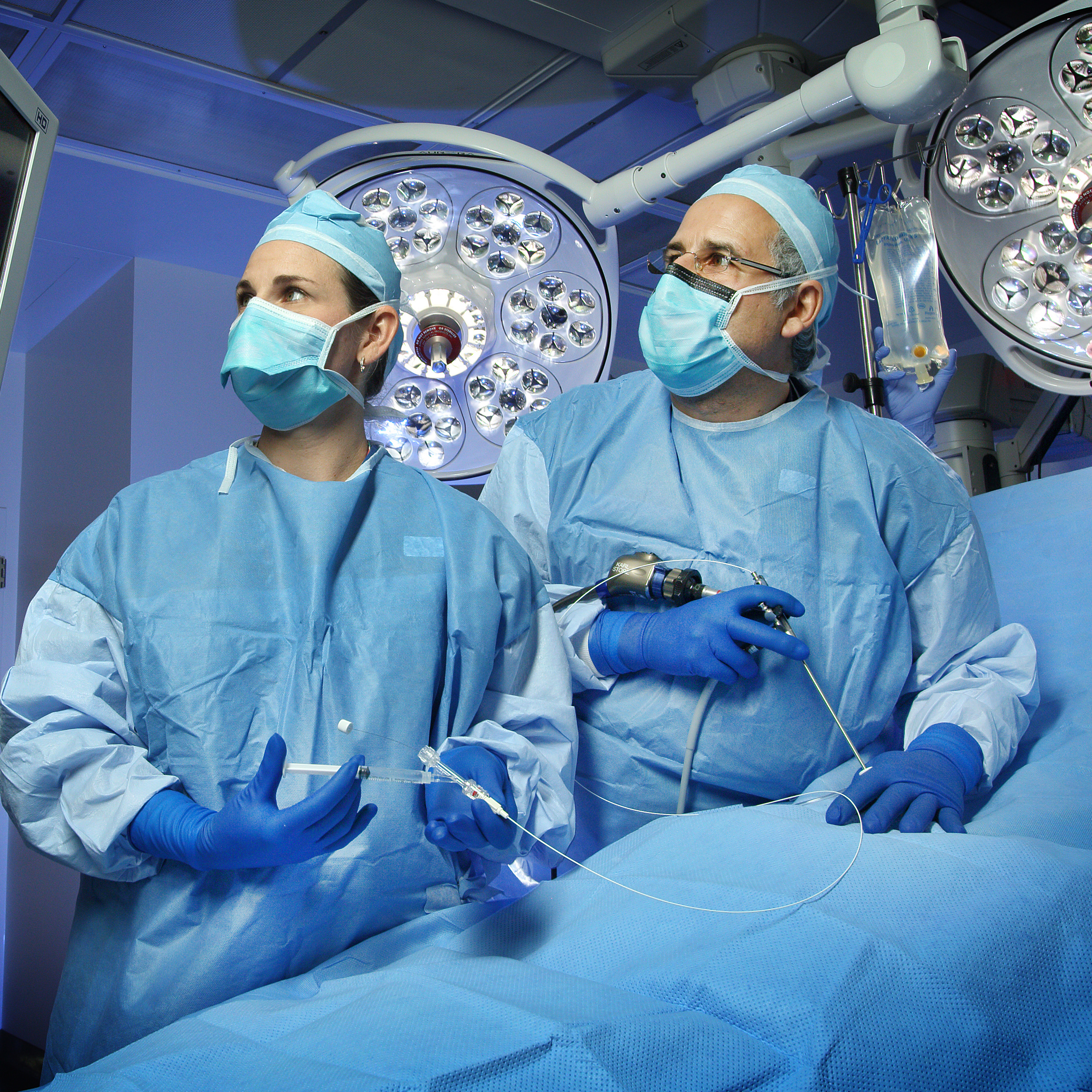 Jena Miller and Ahmet Baschat demonstrate a fetoscope in an OR setting. Both are wearing surgical gowns, caps, masks and gloves and are looking at a monitor screen.