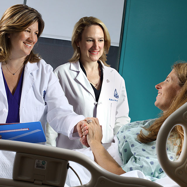 Amanda Fader and Rebecca Stone, wearing white lab coats, smile with a patient laying in a hospital bed as Dr. Fader holds the patients hand.