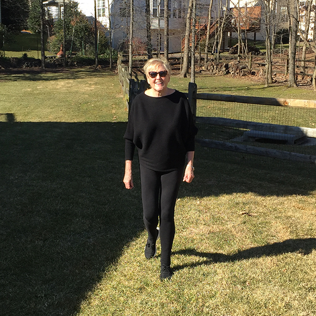 Joan Kravets smiling and walking on grass in a suburban neighborhood with a fence and houses in the background.