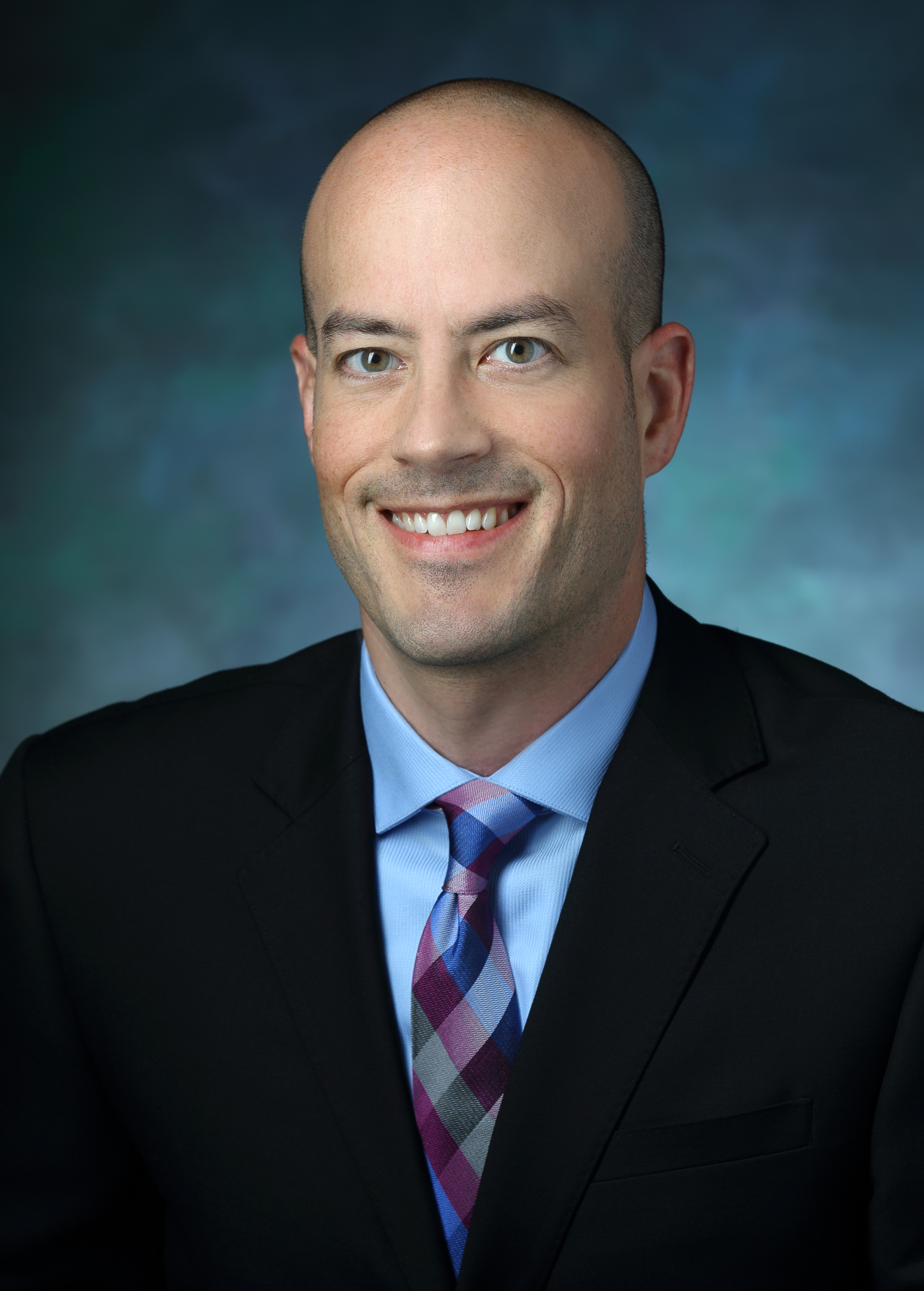 Portrait photo of Ben Larman. He is wearing a black suit with a light blue shirt and a pink, light blue and royal blue plaid tie. He is seated against a blue multicolor background.
