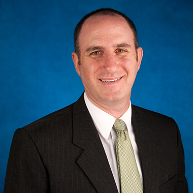 Epidemiologist Eili Klein smiling in a formal portrait with a blue background, wearing a dark-colored suit, beige tie and white button-down shirt.