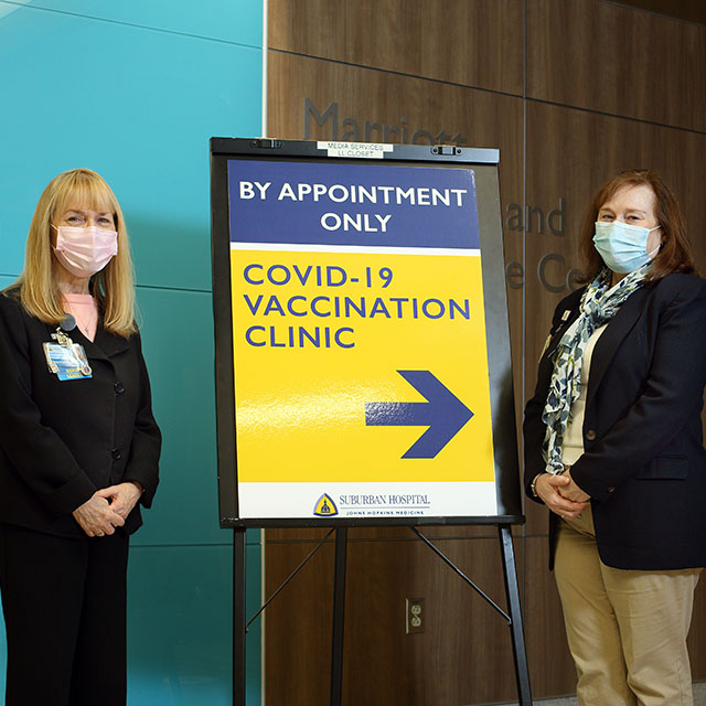 Cindy Notobartolo (left) and Leslie Ford Weber at Suburban Hospital’s community vaccine clinic, standing on either side of a sign that reads: By appointment only. Covid-19 vaccination clinic. 