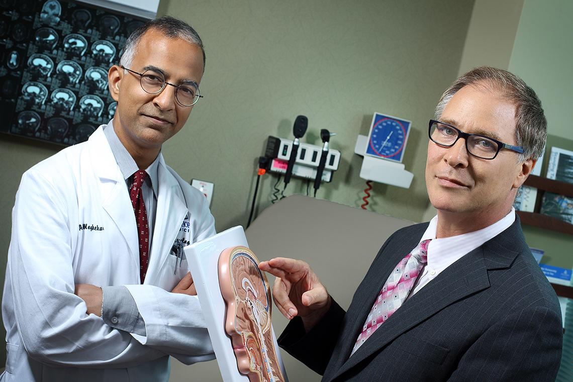 Abhay Moghekar, dressed in a white lab coat, and Mark Luciano, wearing a dark blue suit, holding a 3D model of the brain and neck.