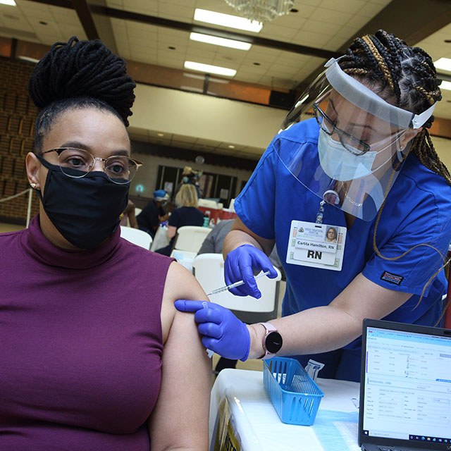 Jetta Ambersley, wearing a purple sleeveless shirt and black mask, gets a COVID-19 vaccine from Hopkins nurse Carlita Hamilton, wearing blue scrubs, a mask and a face shield, at a March clinic in Our Lady of Perpetual Help Roman Catholic Church in Washington, D.C. 