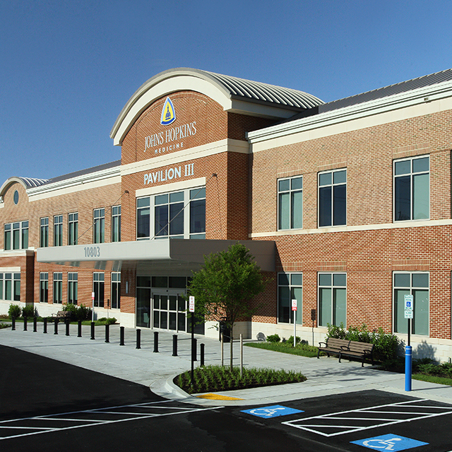 An exterior photo of Pavilion III at Johns Hopkins Health Care & Surgery Center — Green Spring Station shows a brick building with an awning over the entrance, and several parking spaces.