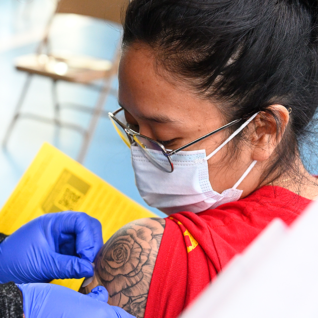 Close-up of a woman’s shoulder, with a tattoo of a rose, as a medical assistant applies a bandage following vaccination. The woman wears a face mask; the medical assistant has on blue surgical gloves, a mask and a face shield. 