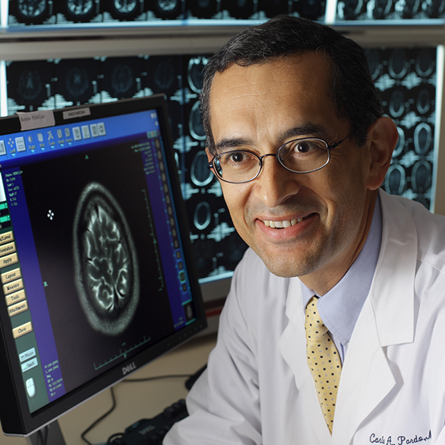 Carlos Pardo-Villamizar wearing a white lab coat in a lab with brain images on screens behind him.