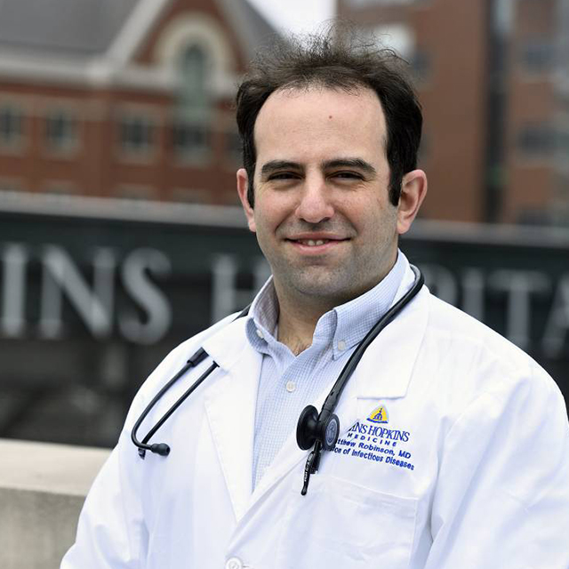 Matthew Robinson, in a white coat with a stethoscope around his neck, with The Johns Hopkins Hospital sign in the background. 