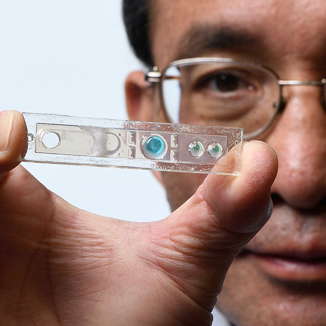 Detail photograph of Tza-Huei Wang holding a tiny device showing small droplets of fluid.