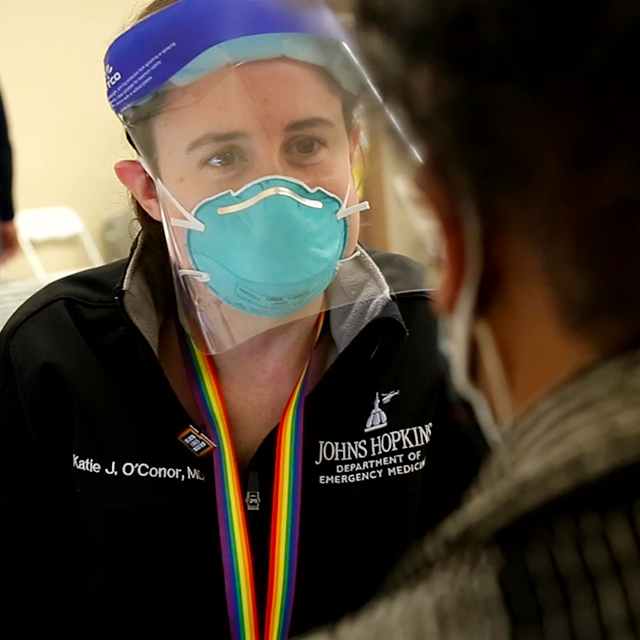 Katie O’Conor, wearing a mask, shield and black jacket that says Johns Hopkins, looking at someone who is not facing the camera.
