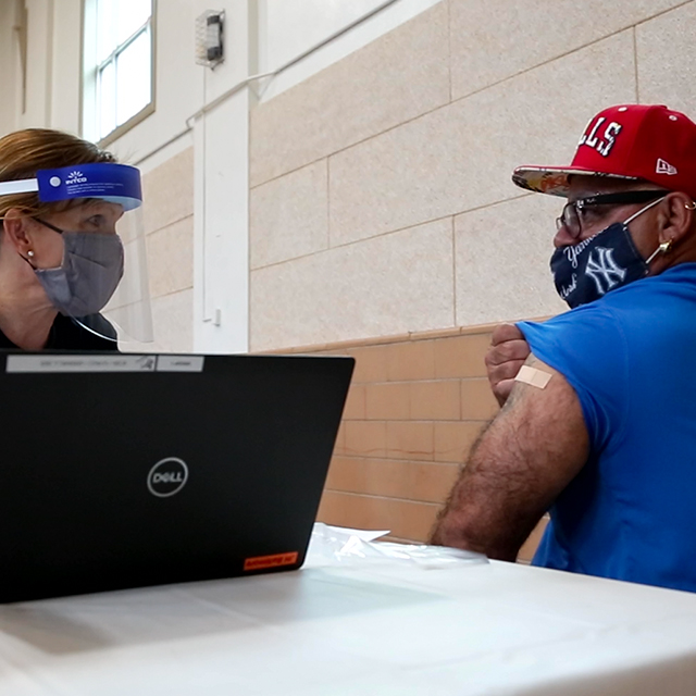 Kathleen Page, wearing a mask and shield, is at a laptop computer next to a man who is lifting his sleeve to show the bandage where he was vaccinated. 
