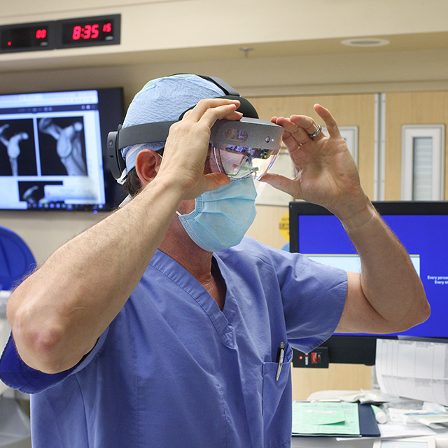 Dr. Rudzki, in scrubs and a surgical mask, wearing augmented reality goggles. A screen in the background shows two images of shoulder ball and socket. 