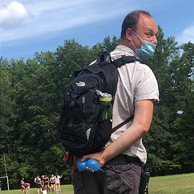 Zilbermint, wearing a mask and a backpack, holding two water balloons behind his back and looking mischievous 