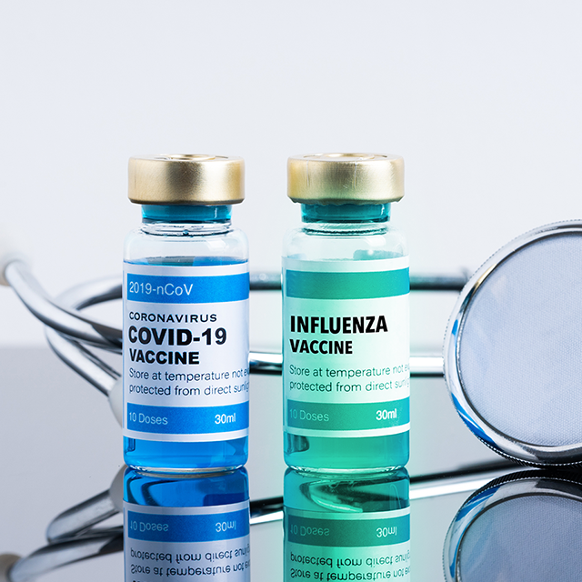 Two vials, one labeled COVID-19 vaccine and one labeled influenza vaccine, sit on a mirrored surface, with a stethoscope behind them.