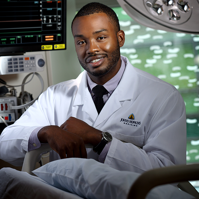 Jason Vaught, wearing a white lab coat, poses in a patient room, leaning on an empty bed, with monitors and a large light behind him.