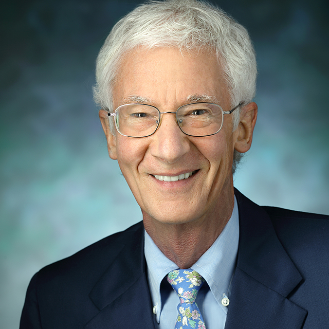 Roland Griffiths smiling in a formal portrait silver-rimmed glasses, a navy blue suit, light blue shirt and flower-print tie