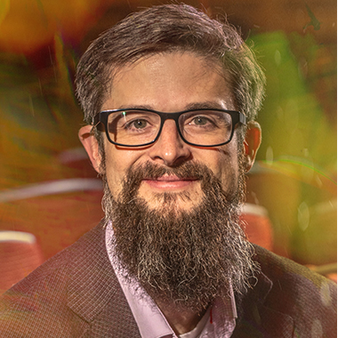 Matthew Johnson, in an altered portrait, wearing black rimmed glasses and a long beard. The image has yellow and orange opaque layers in it.