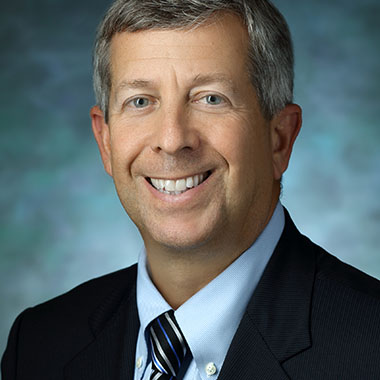 Andrew Satin, in a formal portrait, wearing a blue, silver and black striped tie, light blue button down shirt and dark blue suit jacket