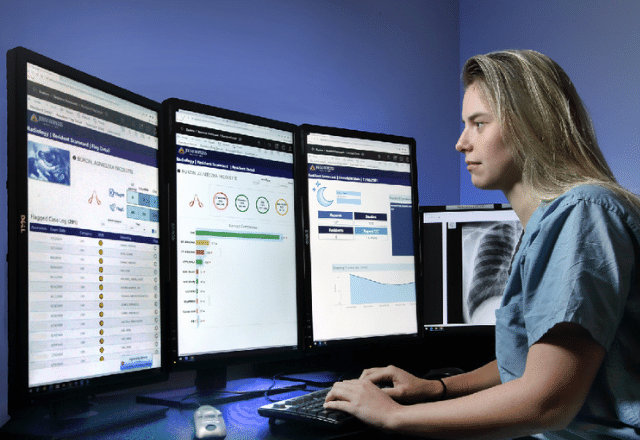 radiologist sitting at a table, reading the dashboard on three computer screens