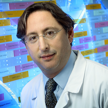 Dorry Segev, wearing a white lab coat, light blue shirt and tie, in front of a multicolored wall displaying the names of organ donors